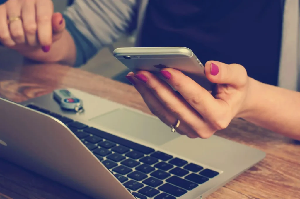 In the picture, a close-up of the hands of a woman who is working on her laptop. In one hand she holds a mobile phone.